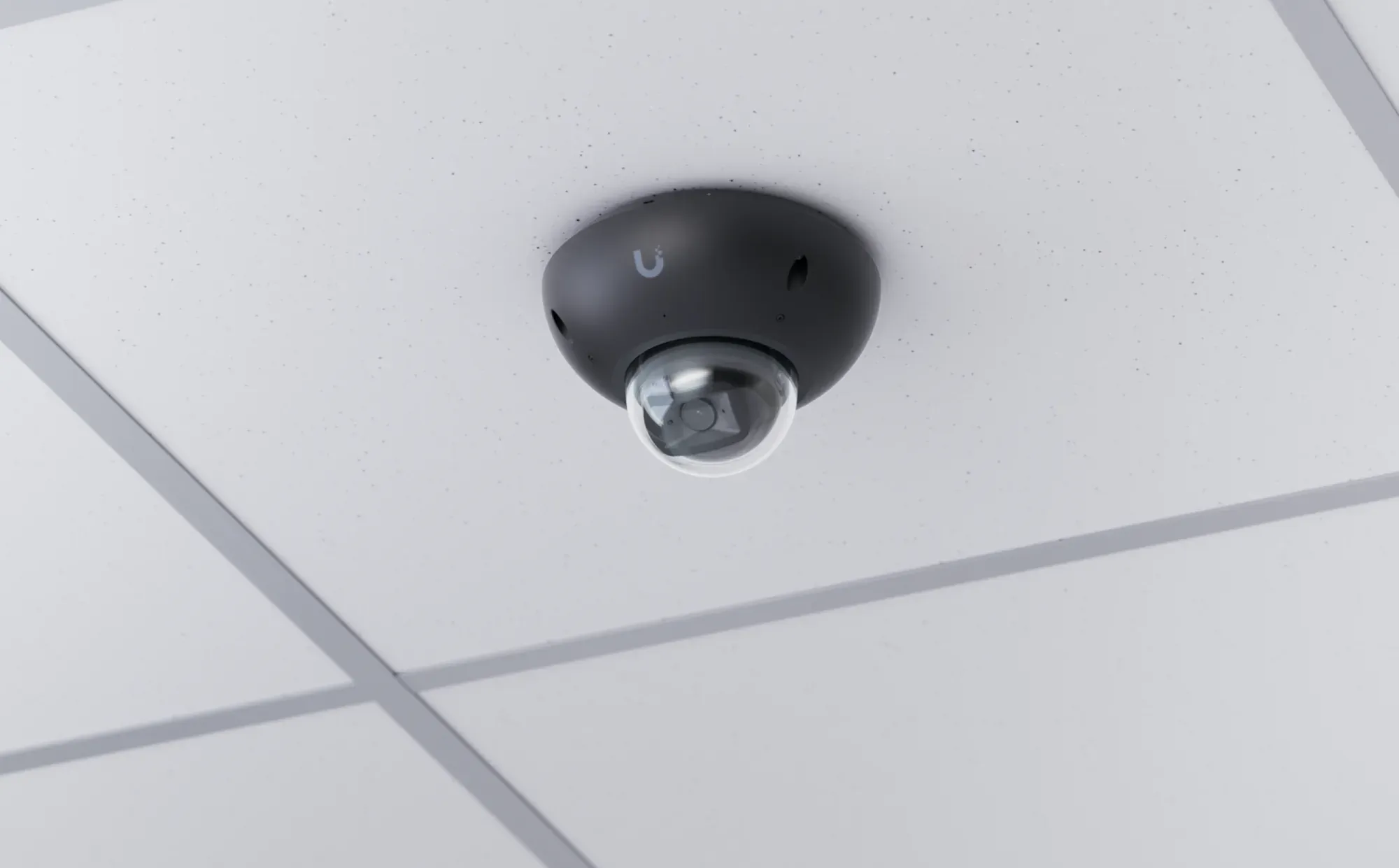 A dome camera on a suspended tile ceiling indoors, seen from below with the lens visible in its clear bubble