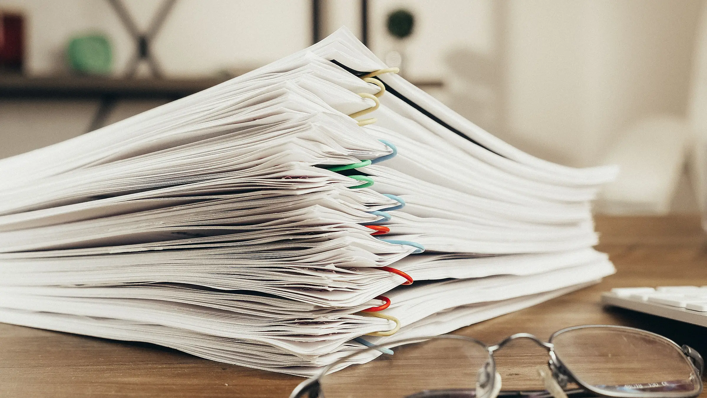 Stack of white paper documents secured with colorful paper clips on a wooden desk next to eyeglasses.