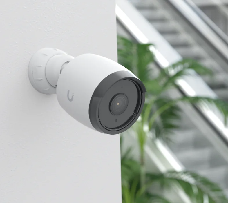 A bullet camera on the outside corner of a rendered wall in daylight, a palm and a glazed stairwell behind it