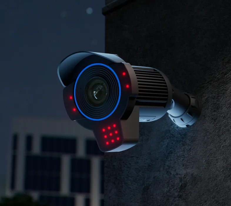 A camera on a rough concrete wall after dark, its infrared emitters lit and a lit office block far behind